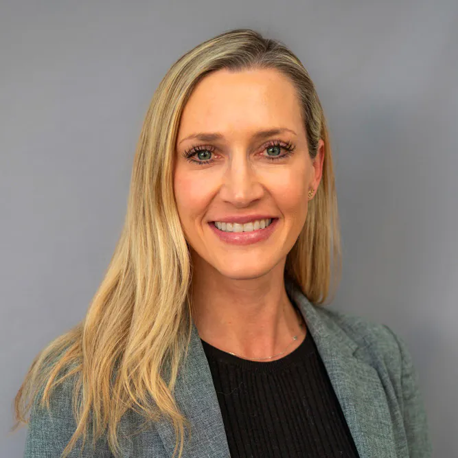 A woman with long blonde hair, wearing a grey blazer over a black top, smiles at the camera against a plain grey background.