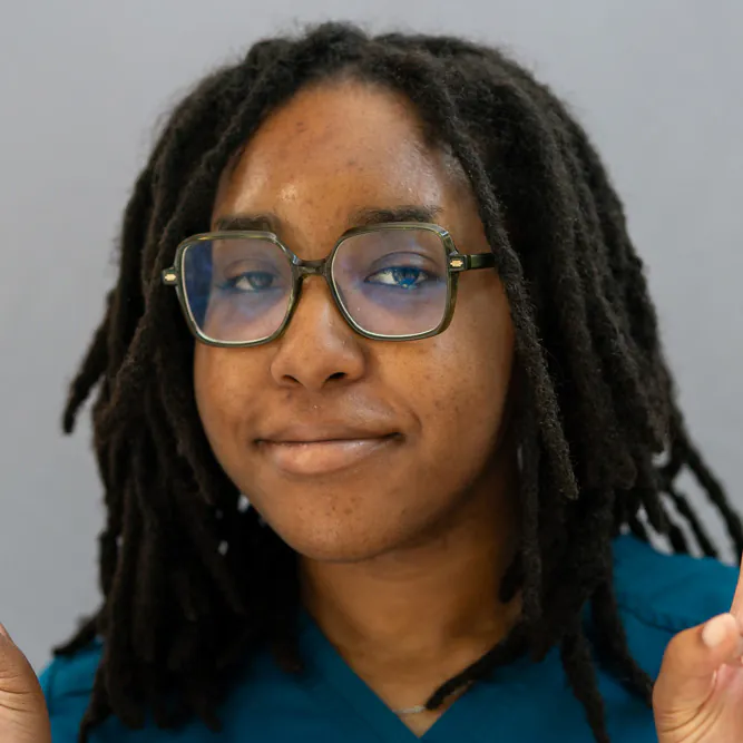 A person with medium-length dreadlocks, wearing glasses and blue scrubs, smiles softly while looking at the camera. The background is plain and gray.