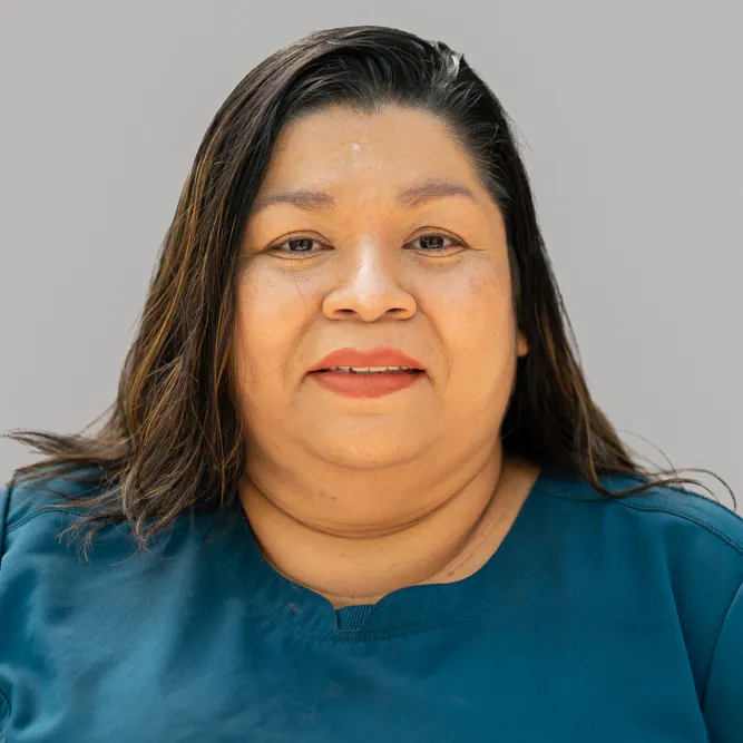 A woman with long dark hair wearing a teal top smiles softly at the camera against a plain light gray background.