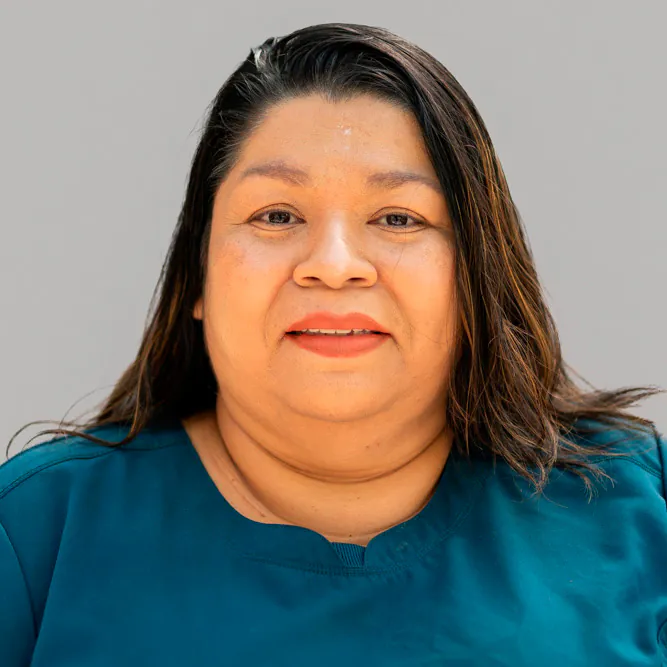 A woman with long dark hair, wearing a teal shirt, is looking at the camera and smiling softly. The background is plain and light gray.