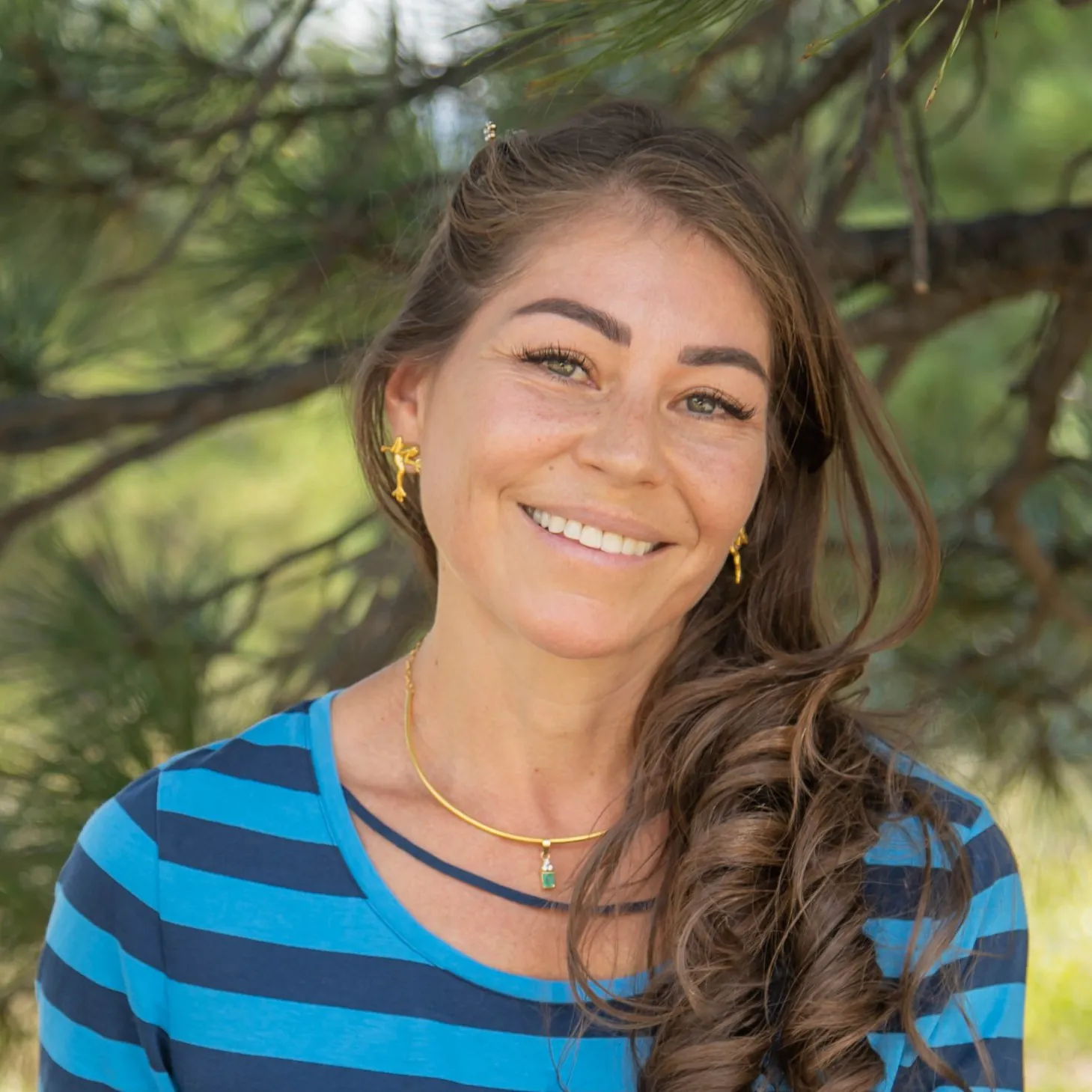 A woman with long brown hair wearing a blue striped shirt and gold jewelry smiles while standing outdoors in front of pine tree branches.