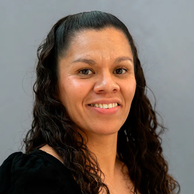 A woman with long, curly brown hair and a black top smiles at the camera against a plain gray background.