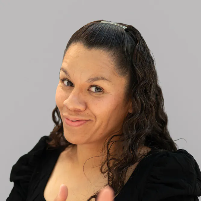 A woman with long, wavy brown hair and a hair clip smiles at the camera. She is wearing a black top and is posed against a plain, light gray background.