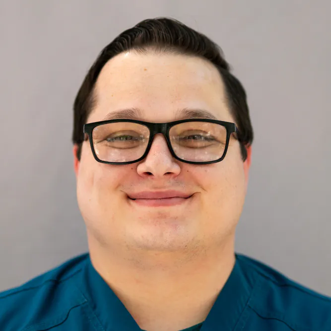 A person with short dark hair and black glasses smiles at the camera, wearing a teal shirt against a plain light gray background.