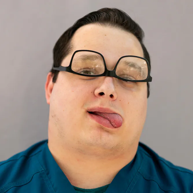 A man in teal scrubs with glasses askew on his face sticks out his tongue and looks at the camera against a plain gray background.