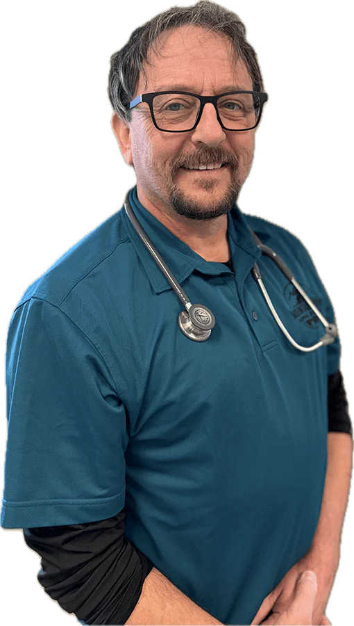 A man wearing glasses and a teal polo shirt with a stethoscope around his neck smiles at the camera.