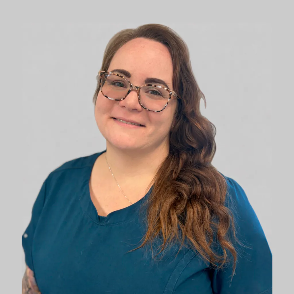 A woman with long wavy brown hair, wearing glasses and a blue top, smiles at the camera against a plain light gray background.