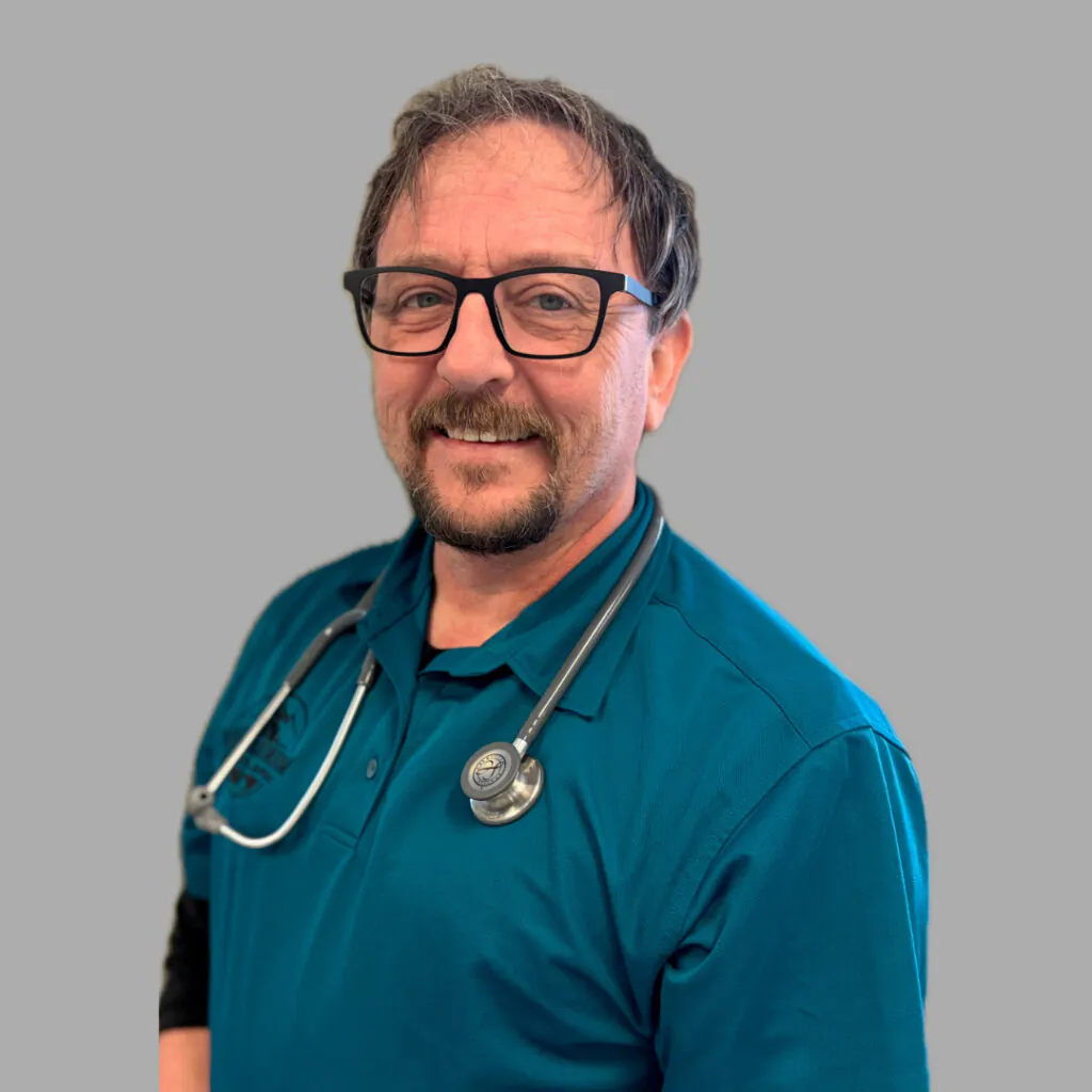 A middle-aged man with glasses, a beard, and mustache smiles while wearing a teal polo shirt and a stethoscope around his neck, standing against a plain gray background.