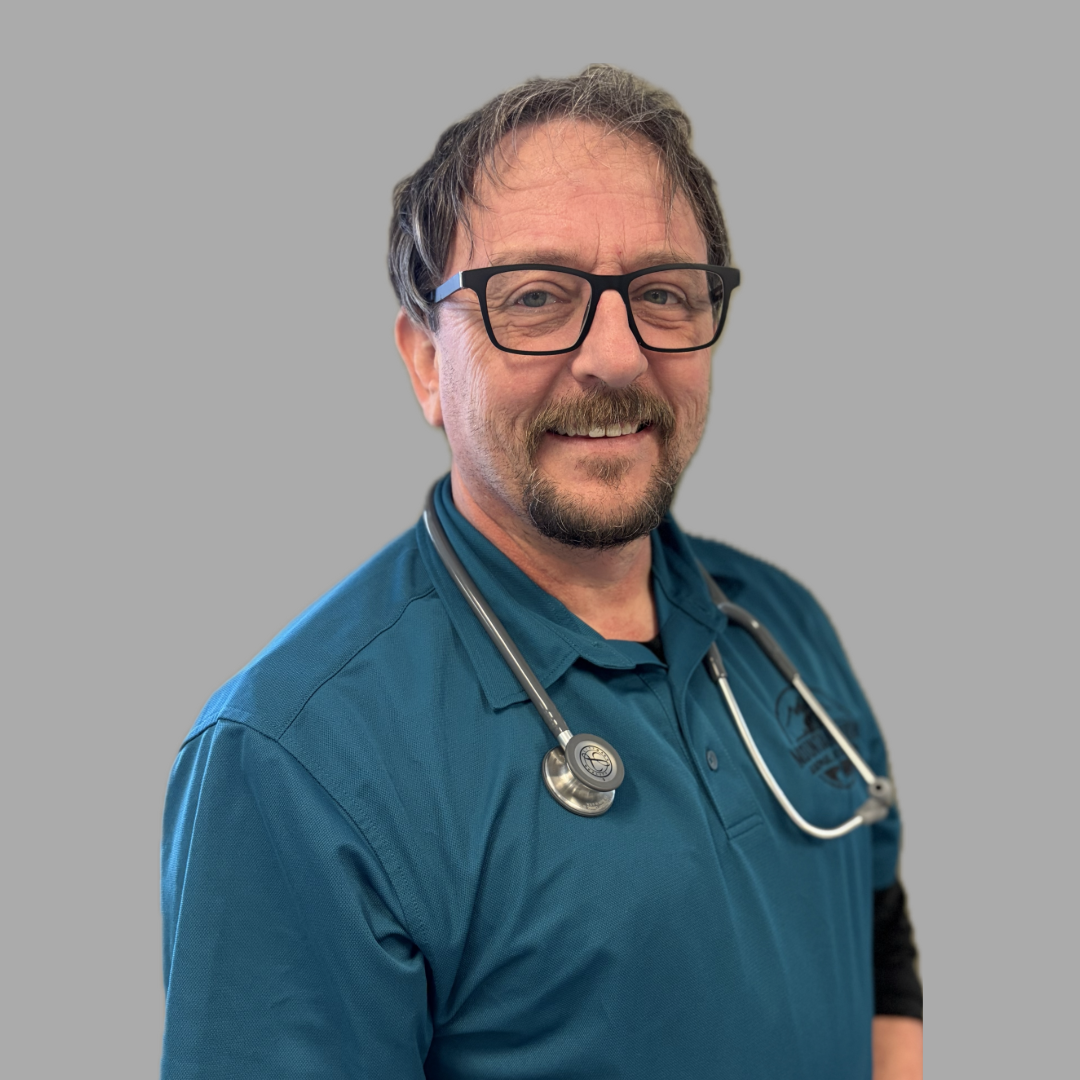 A smiling man wearing glasses, a teal collared shirt, and a stethoscope around his neck stands in front of a plain grey background.