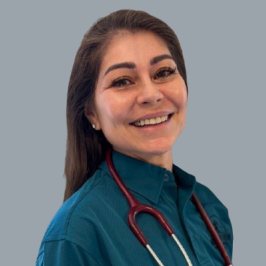 A woman with long brown hair, wearing a teal shirt and a stethoscope around her neck, smiles at the camera against a plain gray background.