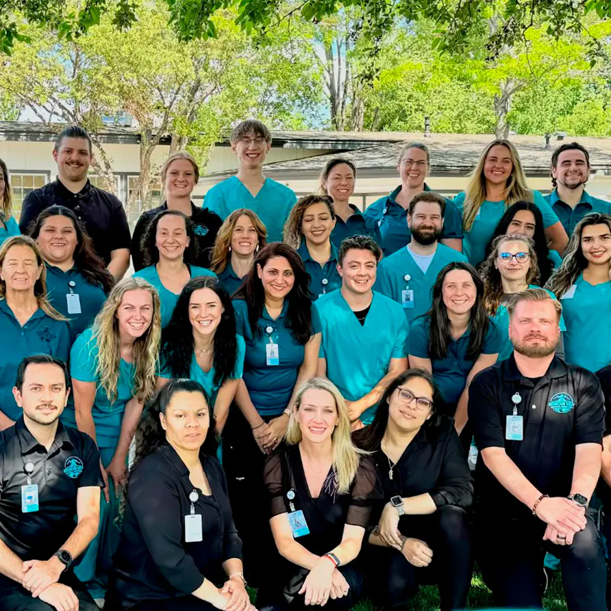 A group of healthcare professionals in teal and black uniforms pose outdoors under trees, smiling at the camera. There are about 26 people, with name badges, in three rows on a sunny day.