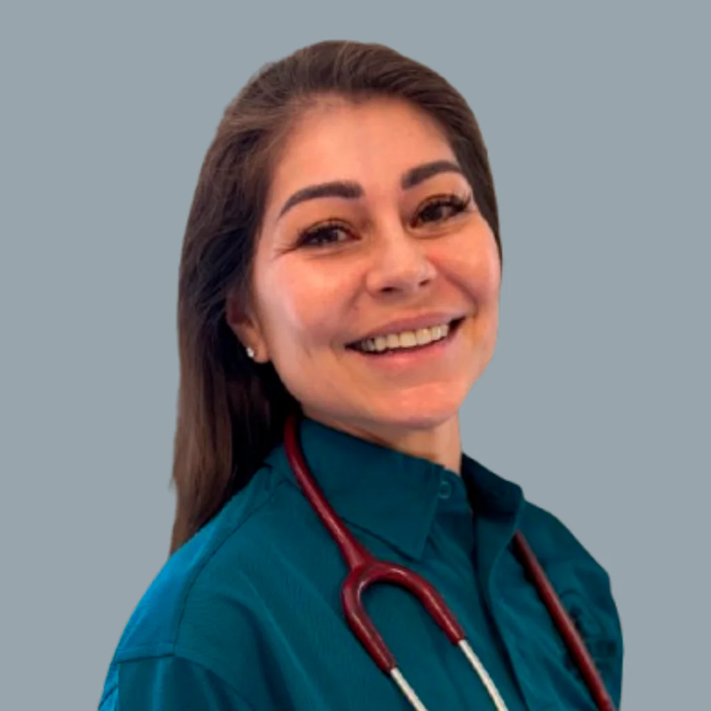 A woman with long brown hair, wearing a teal shirt and a stethoscope around her neck, smiles at the camera against a plain gray background.